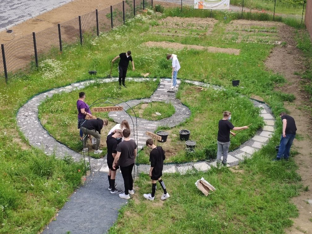 Das Bild zeigt mehrere Jugendliche aus der Vogel-Perspektive, die einen Betonkreis in einer Wiese im Weltethos Schulgarten in Merzenich bauen.