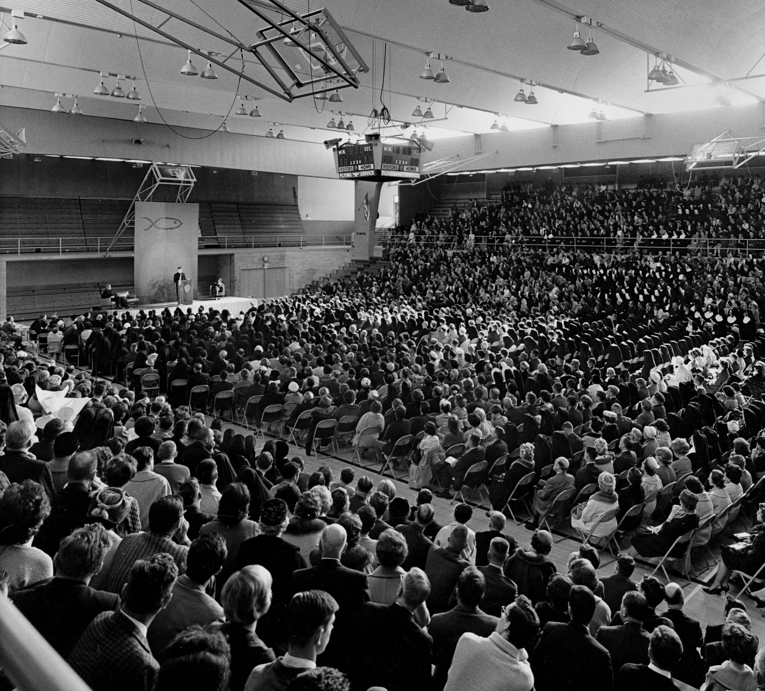 Das Bild (schwarz-weiß) zeigt Hans Küng, den späteren Gründer der Stiftung Weltethos, bei einer Rede vor einer Turnhalle voll Menschen auf Stühlen in San Francisco.