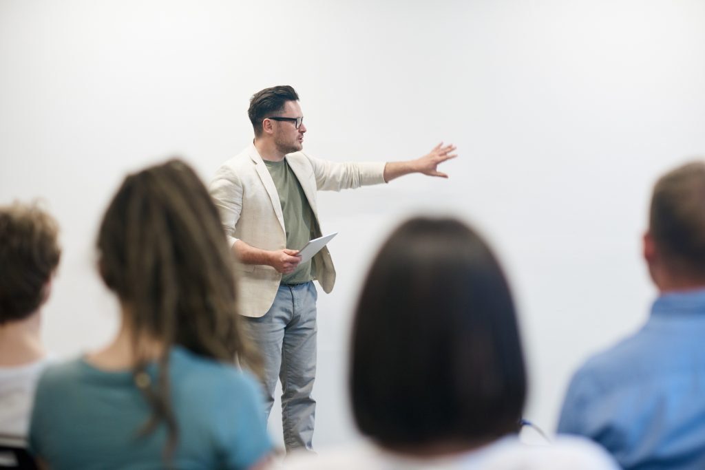 Das Bild zeigt einen jungen Mann. Er steht vor einer Klasse und hält einen Vortrag.