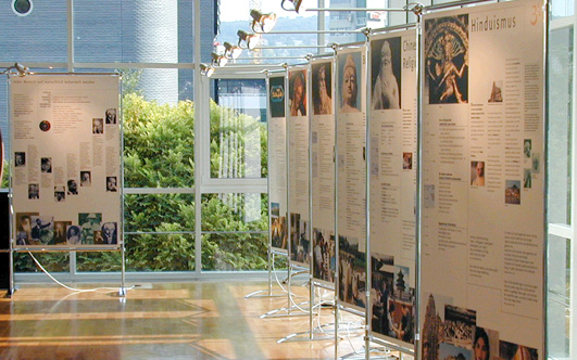 Das Bild zeigt sieben große Informationstafeln aufgestellt nebeneinander in einem Stellsystem und mit Beleuchtung als Weltethos Ausstellung Version A.