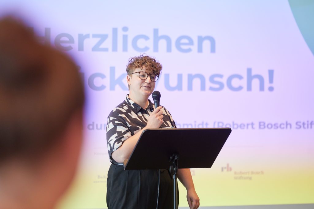 Luisa Schmidt, Projektmanagerin bei der Robert Bosch Stiftung, steht vor einem kleinen Pult und hält die Laudatio für den zweiten Platz. Sie hält ein Mikrofon in der Hand und schaut ins Publikum.