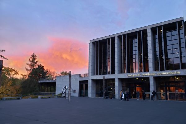 Zu sehen ist ein Gebäude mit der Aufschrift "congress centrum Weimarhalle". Davor ist ein Platz mit Menschen. Der Himmel ist bunt gefärbt.